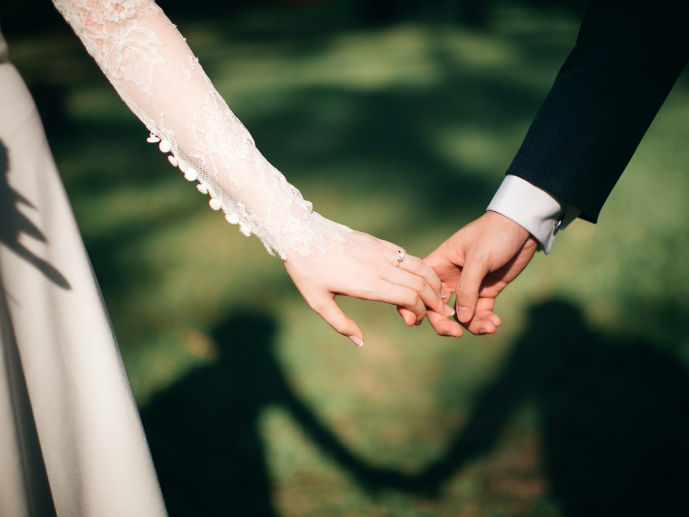 Close view of a bride in a lace sleeve and a groom in a dark suit holding hands, their shadow on the grass