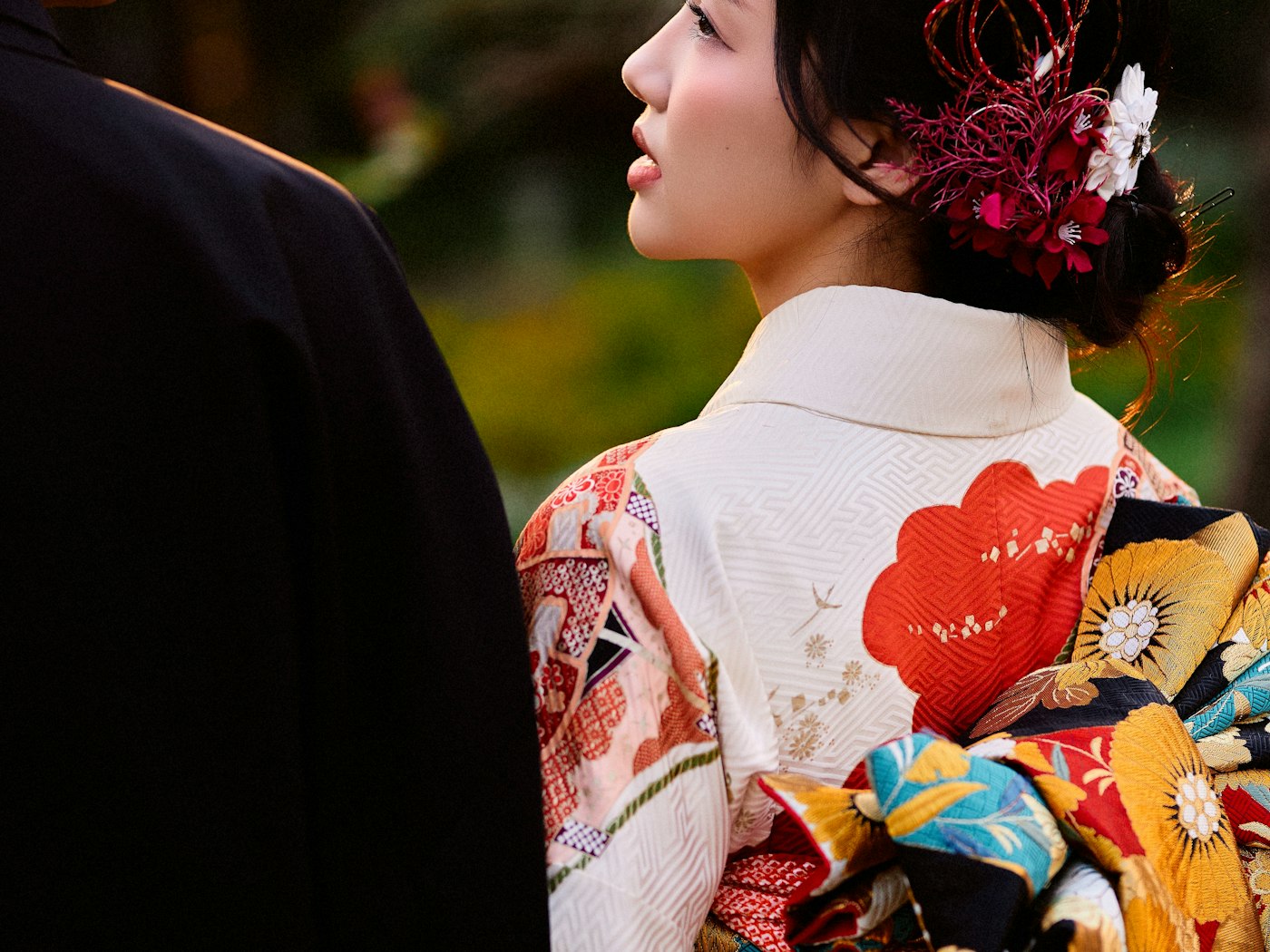 Bride in a patterned white uchikake seen over her shoulder, a red kanzashi ornament in her hair