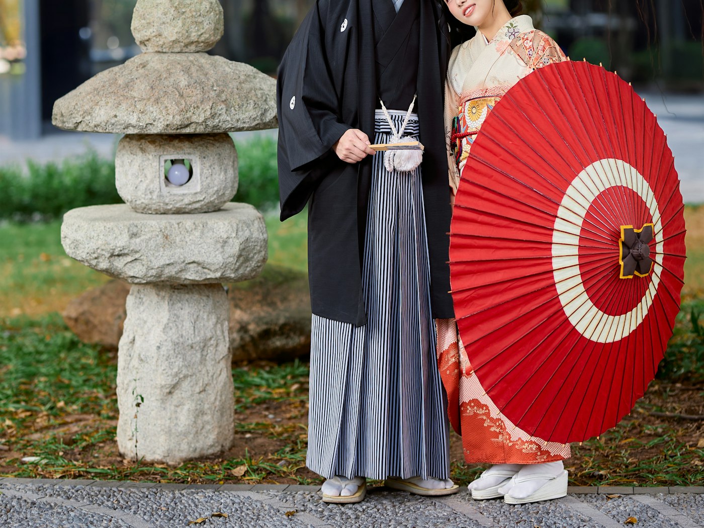Couple in kimono standing beside a stone lantern with a red paper parasol