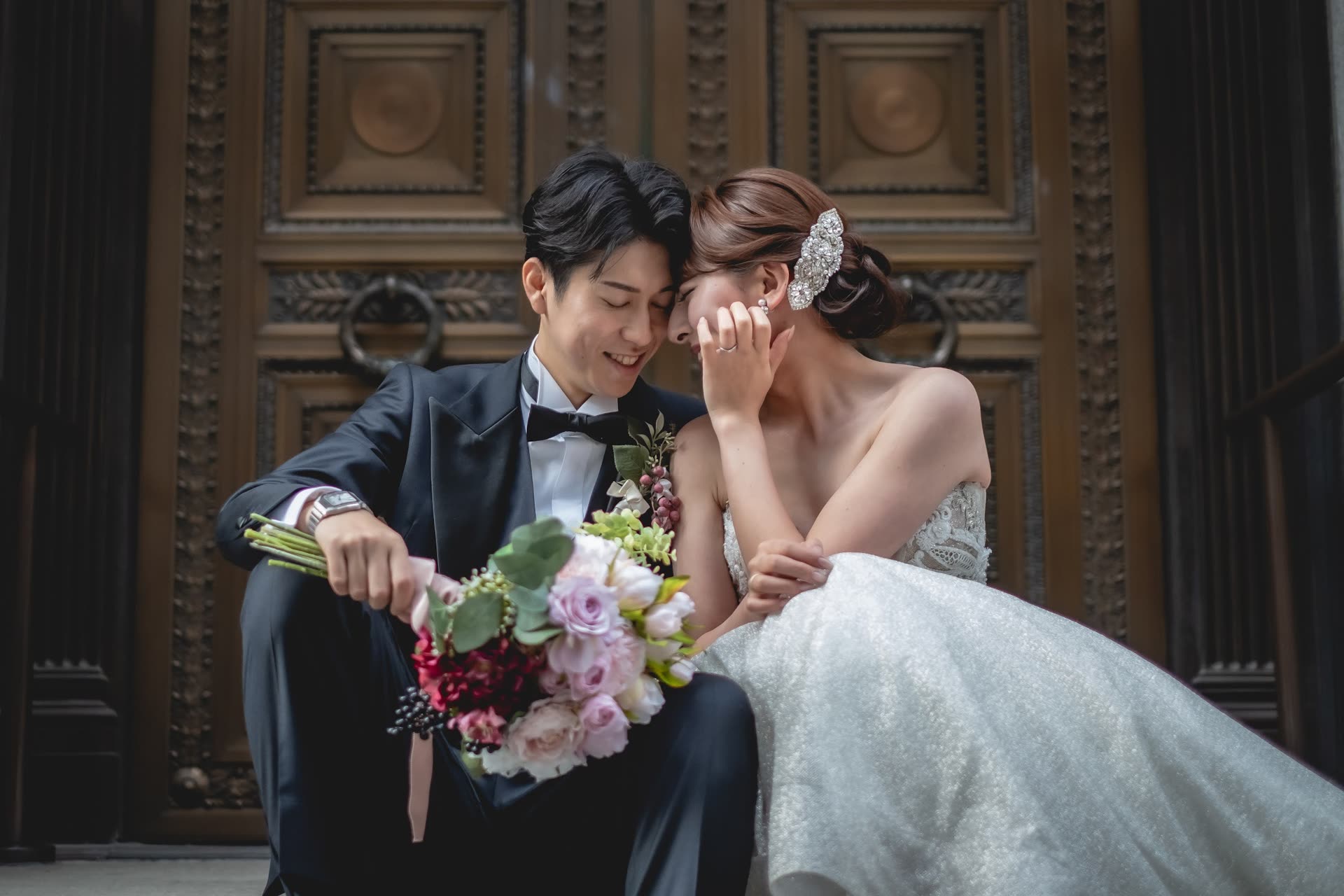 Couple seated on steps before heavy heritage doors, the bride holding a garden bouquet