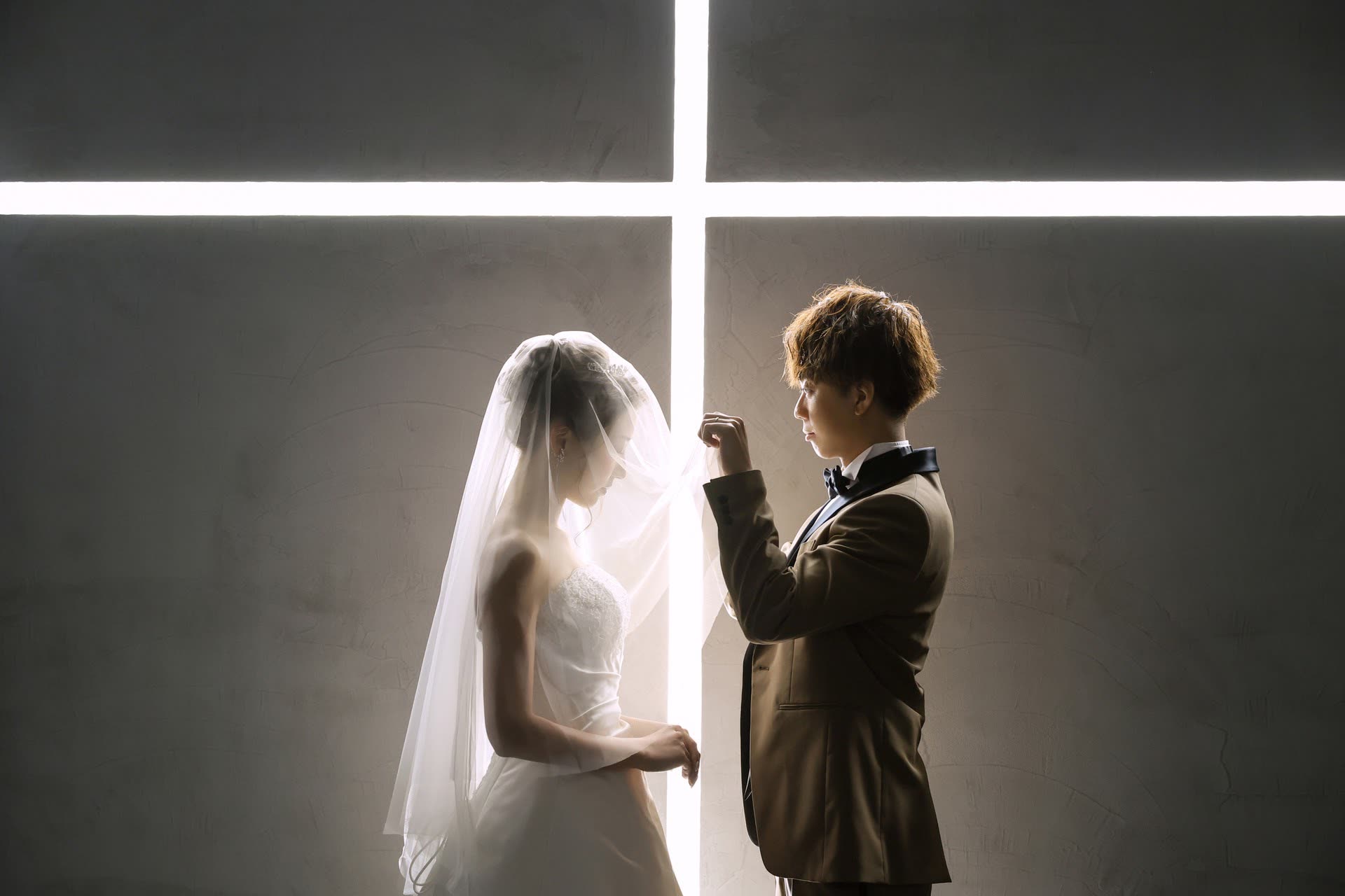 Groom lifting the bride's veil before a cross of light in a modern Japanese chapel