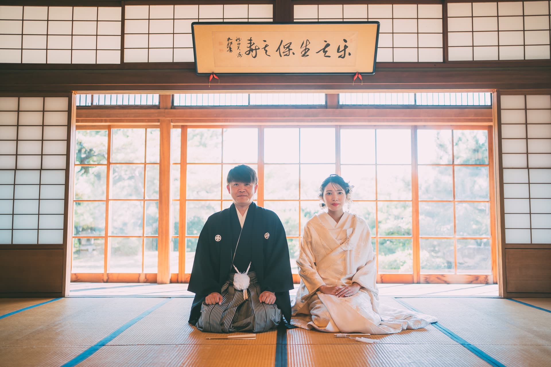 Bride and groom in kimono seated formally on tatami beneath a calligraphy plaque in a Japanese house