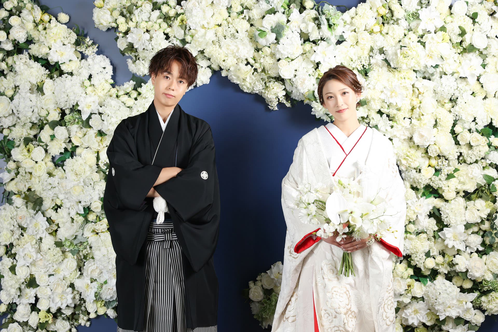 Couple in shiromuku and montsuki-hakama before a wall of white flowers in a Japanese studio
