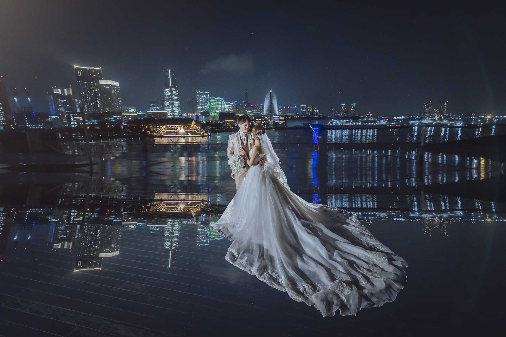 Couple in a long gown and suit beside still water at night, the city lights reflected around them, on a pre-wedding photoshoot in Japan