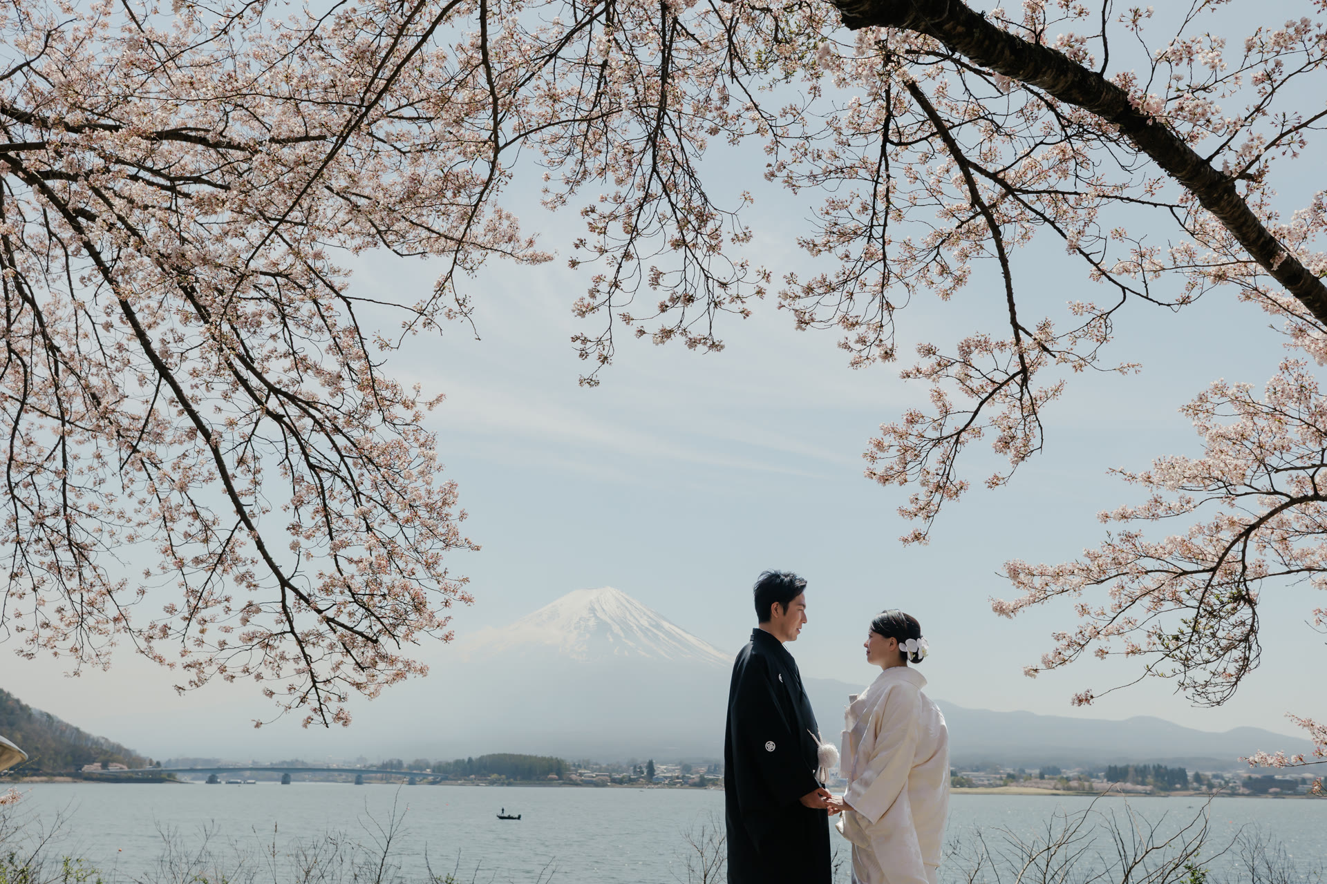 Couple in traditional kimono beneath cherry blossom branches on the shore of Lake Kawaguchi, with snow-capped Mt. Fuji across the water