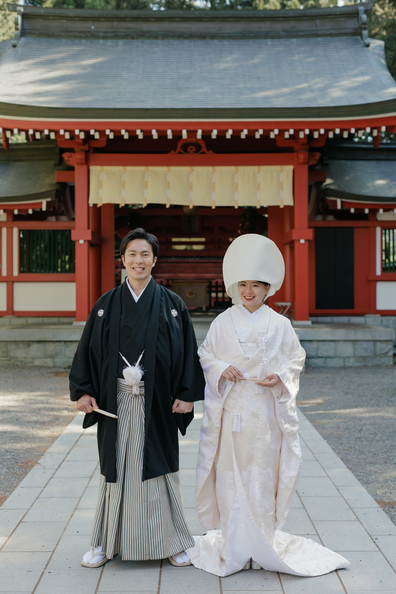 Bride in shiromuku and wataboshi and groom in montsuki standing formally before a shrine hall