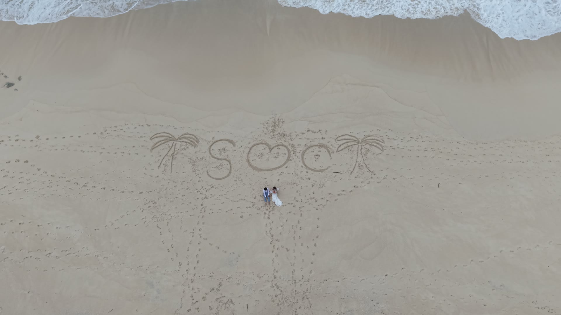 Aerial view of a couple on a white-sand Okinawa beach beside a heart drawn in the sand, on a pre-wedding photoshoot