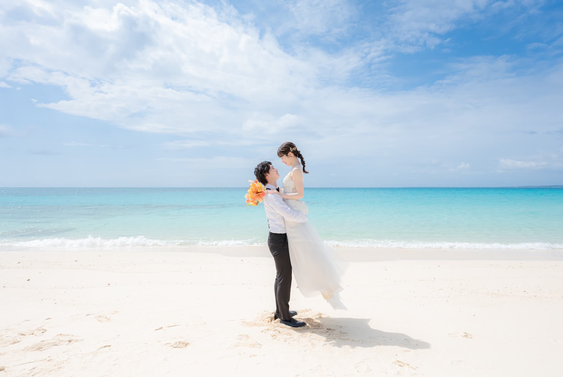 Groom lifting his bride on white sand with the turquoise Okinawa sea behind them