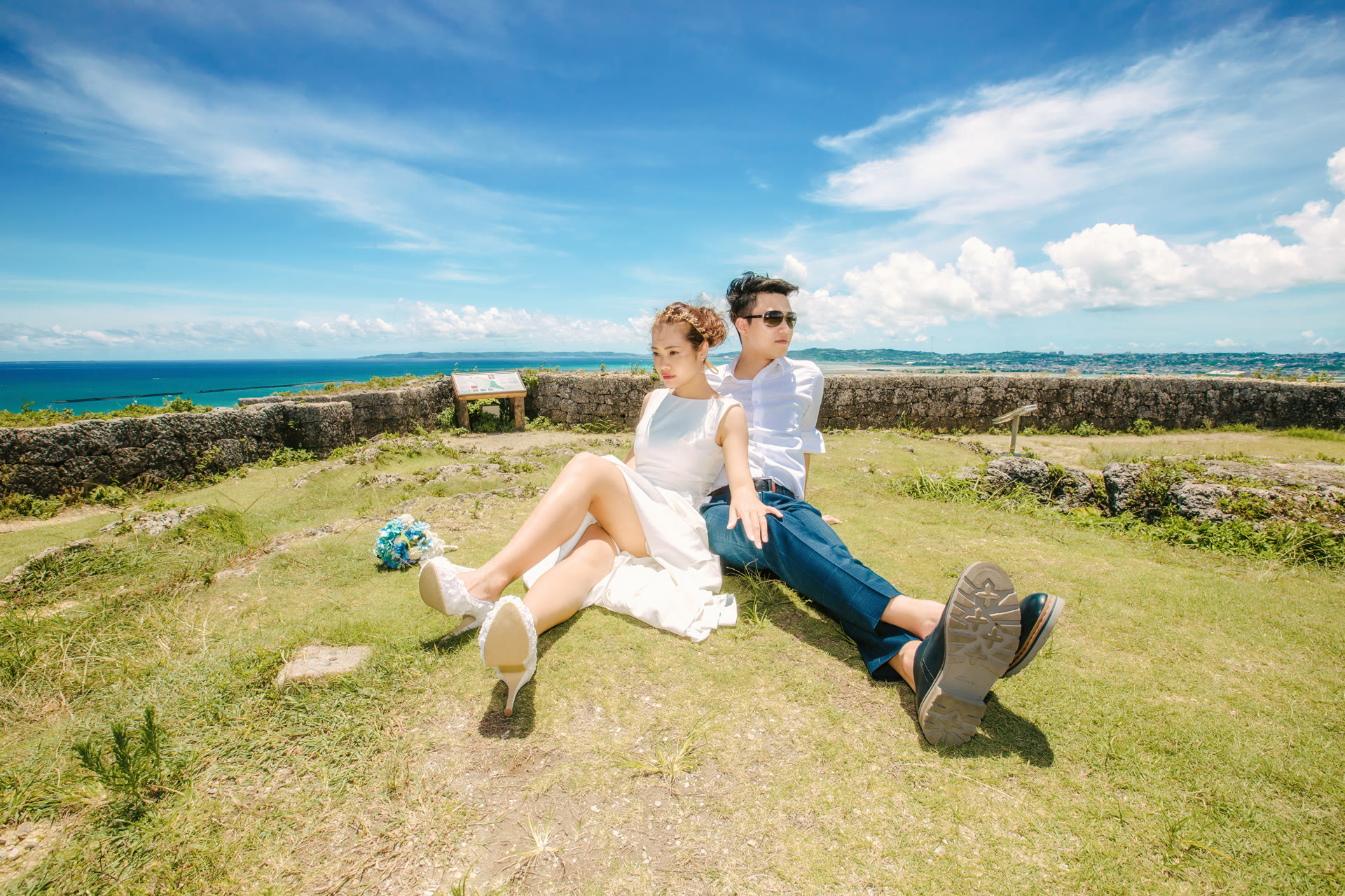 Couple sitting on grass beside Ryukyu stone walls with the ocean behind