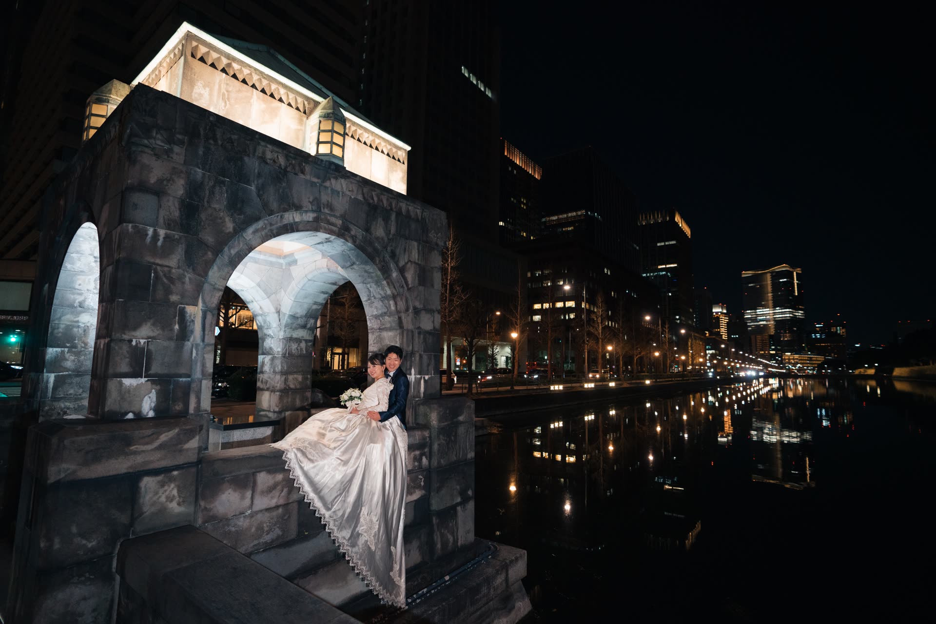 Bride in a long gown seated inside a lit stone arch beside a Tokyo river at night