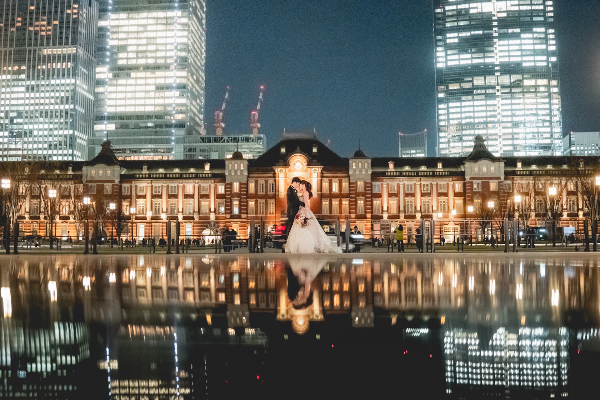 Couple photographed before the illuminated brick facade of Tokyo Station, mirrored in still water at night